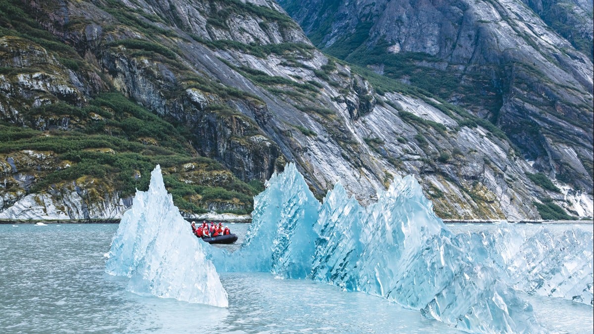 A kayaker paddling in calm, clear waters of an Alaskan fjord with towering green cliffs and a small boat in the distance