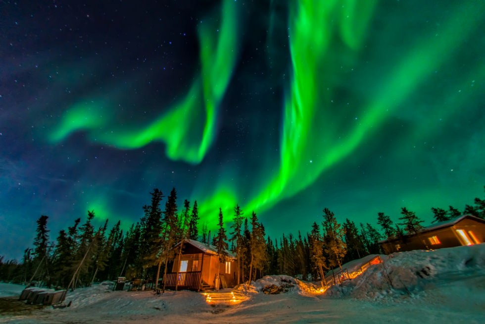 Vivid emerald curtains of Northern Lights dancing over snow-covered wilderness lodge cabin