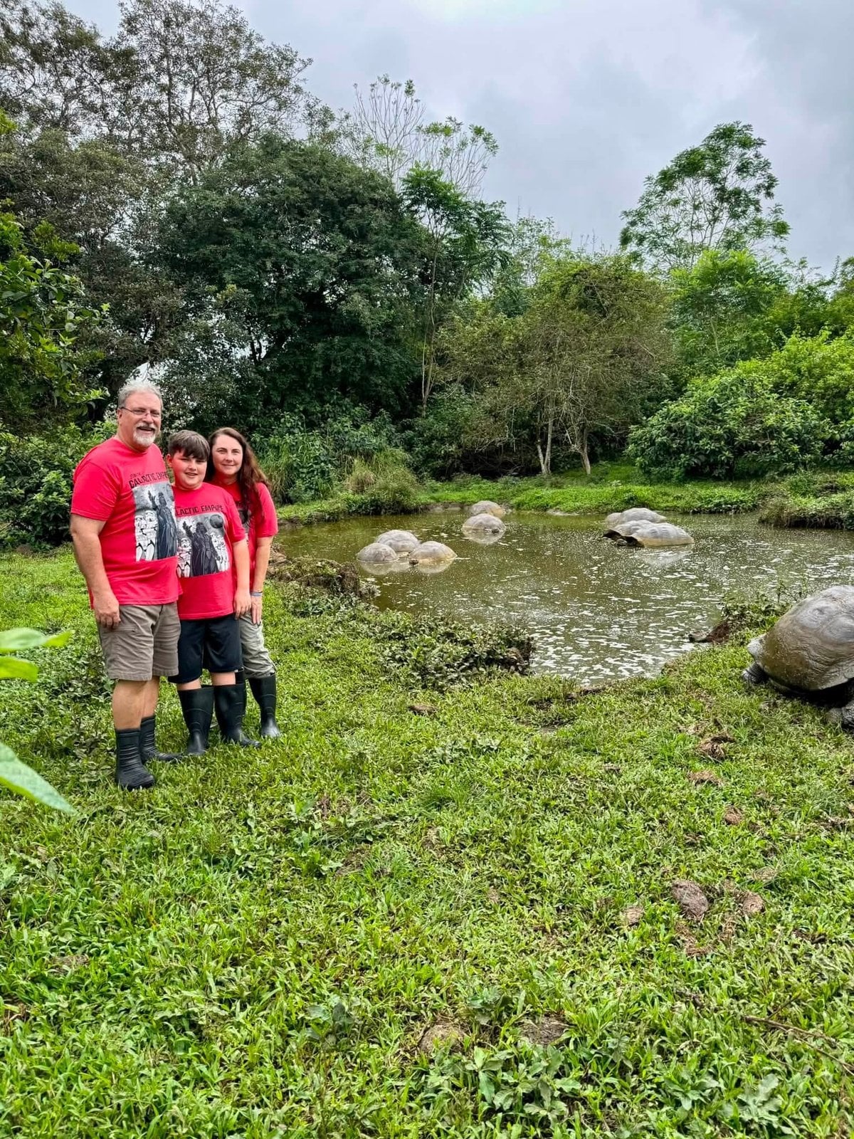 The Herring family in red shirts and rubber boots standing by a freshwater pond with wild Giant Galapagos Tortoises