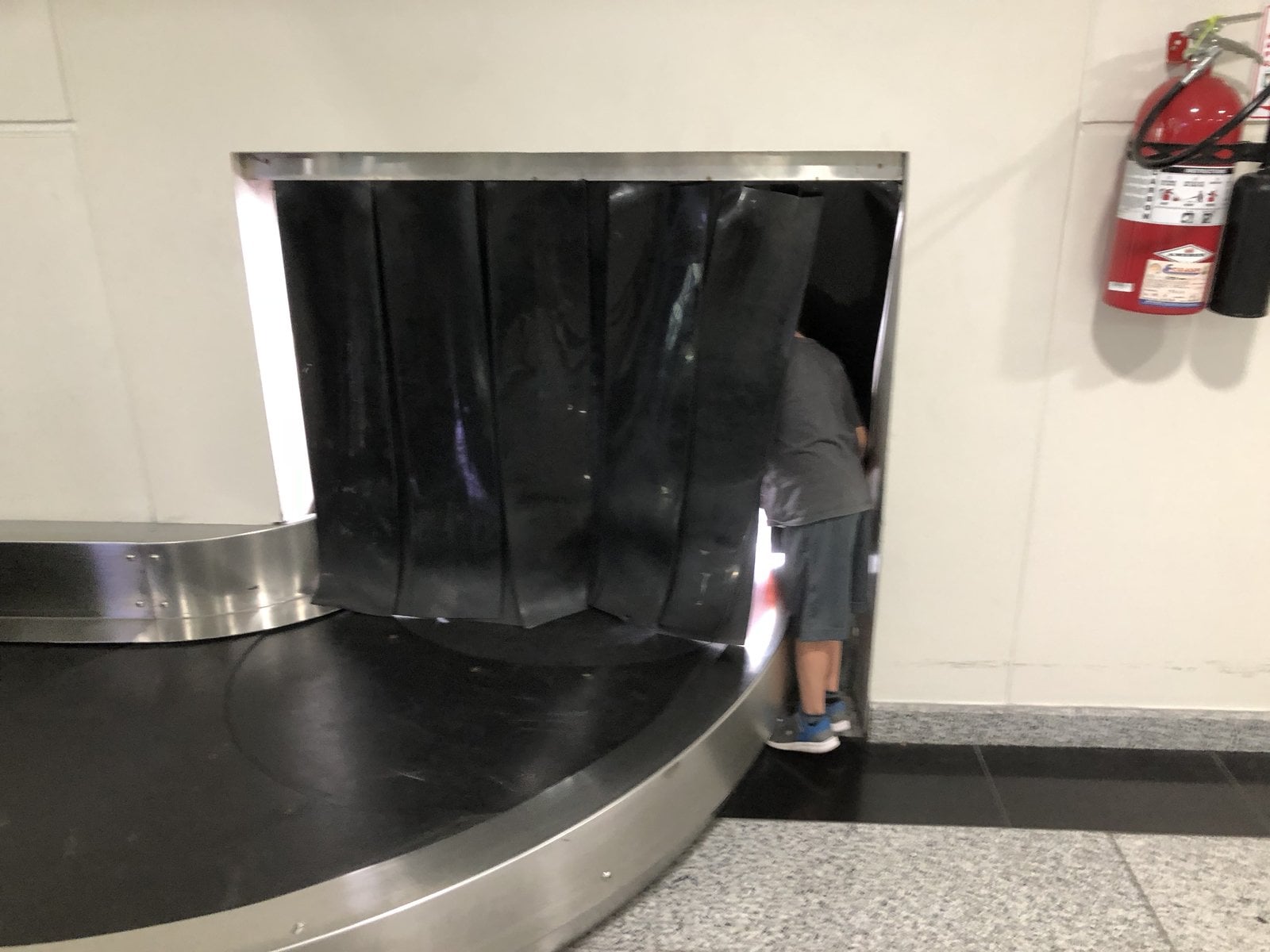 Karl Kannstadter son peering into empty luggage carousel flap at airport